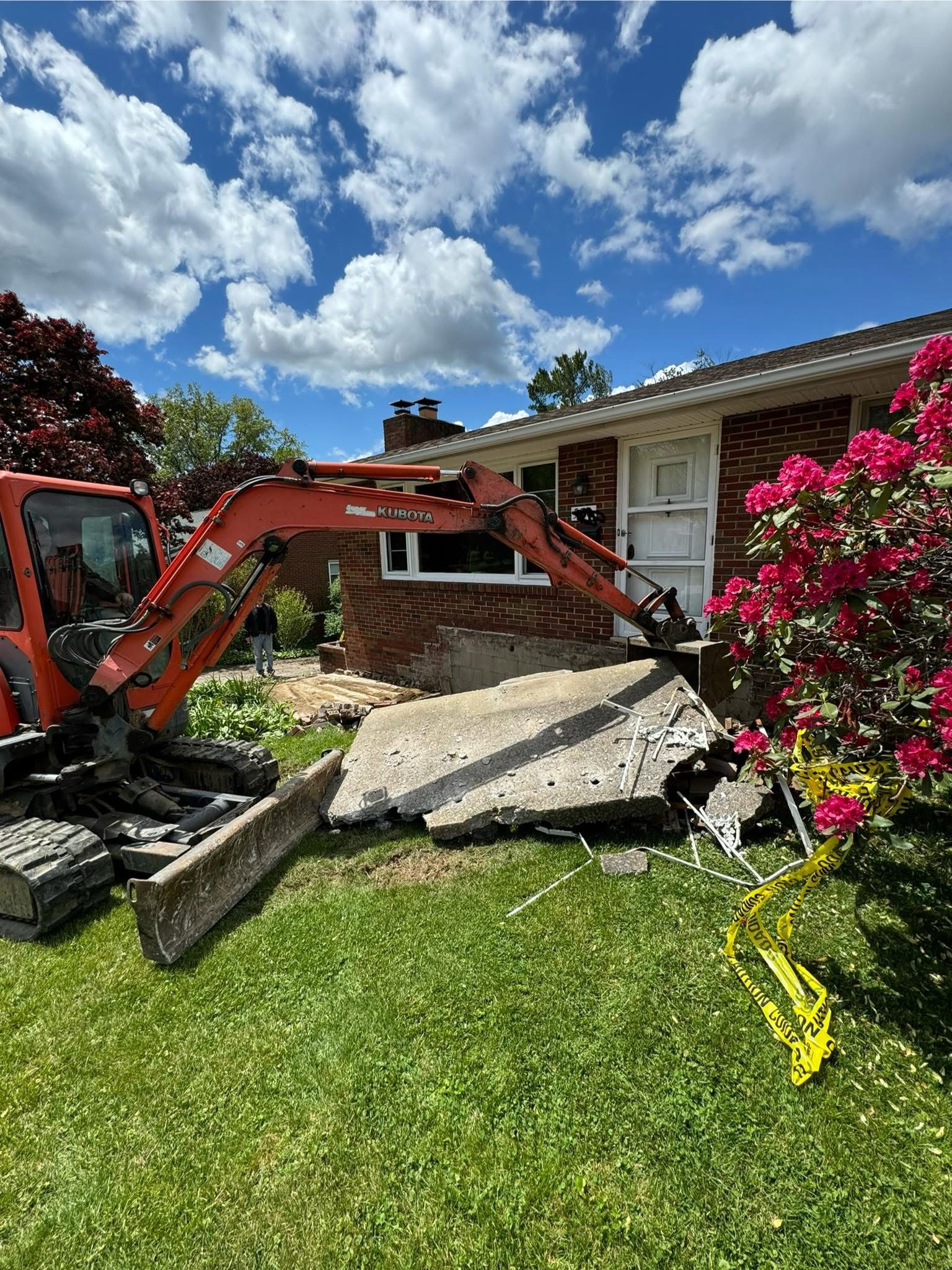 Excavator removing an old concrete slab in front of a brick home